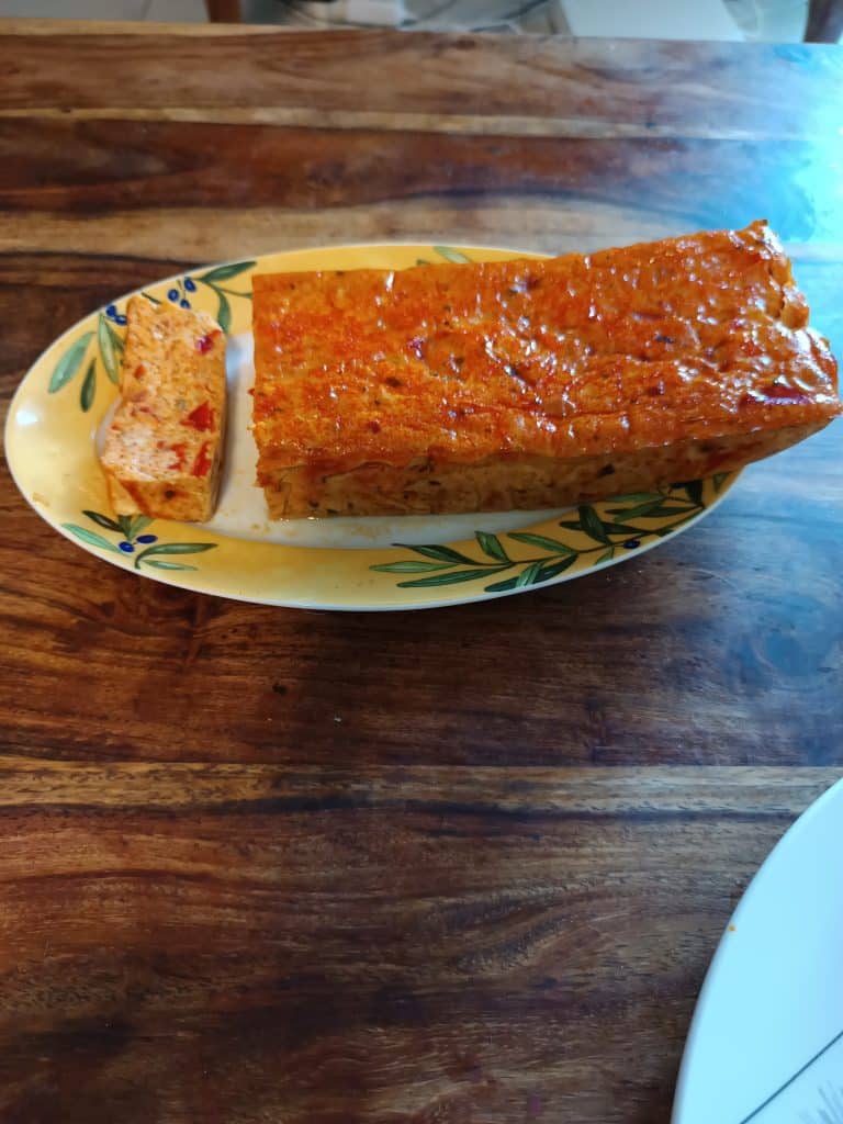 Le pain de tomate, posé dans un plat de service aux couleurs d'été, jaune avec des brins d'olive, et une tranche de ce pain découpée